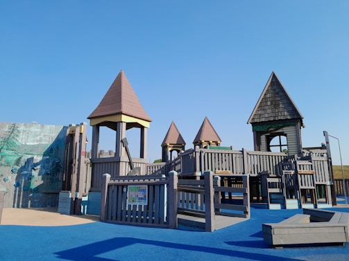 A children's playground with wooden structures resembling small castles and towers. The playground is set on a blue rubber floor with various climbing and play equipment.