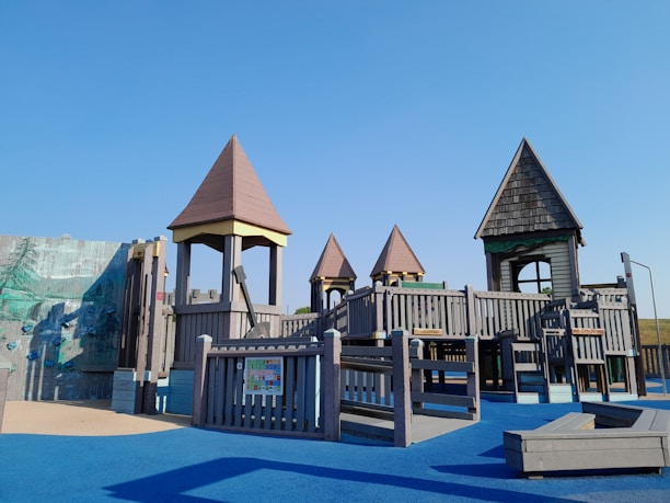 A children's playground with wooden structures resembling small castles and towers. The playground is set on a blue rubber floor with various climbing and play equipment.