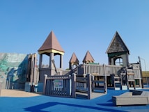 A children's playground with wooden structures resembling small castles and towers. The playground is set on a blue rubber floor with various climbing and play equipment.