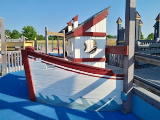 A playground structure designed to look like a ship made of wood with white and red painted sections. It features a circular window and is surrounded by a blue ground surface that mimics water. Wooden fences and additional playground equipment are visible in the background with some trees and grass under a clear blue sky.