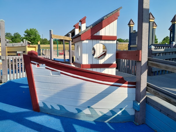 A playground structure designed to look like a ship made of wood with white and red painted sections. It features a circular window and is surrounded by a blue ground surface that mimics water. Wooden fences and additional playground equipment are visible in the background with some trees and grass under a clear blue sky.