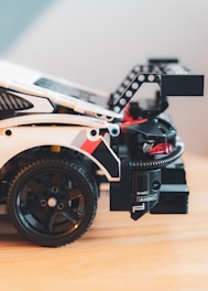 A detailed image of a toy car featuring intricate design elements like a large rear wing, visible wheel with a black rim, and various decals. The car appears to be constructed from interlocking plastic pieces with multiple colors like black, white, and red, giving it a sporty appearance. The scene is set on a wooden surface with a soft, blurred background.