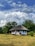 A quaint, traditional house with a thatched roof is set amidst a lush, green landscape. Large clouds fill the blue sky above, adding a dramatic backdrop to the serene scene.