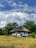 A quaint, traditional house with a thatched roof is set amidst a lush, green landscape. Large clouds fill the blue sky above, adding a dramatic backdrop to the serene scene.