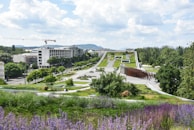 A modern landscape with expansive green spaces, pathways, and architectural elements in the foreground. Trees are spaced along the path leading up to a large, flat building in the distance. The area is lush with greenery and features both vibrant purple flowers and neatly trimmed grass. A few people are visible, enjoying the park area. A construction crane and urban buildings are visible in the background, with hills further beyond.