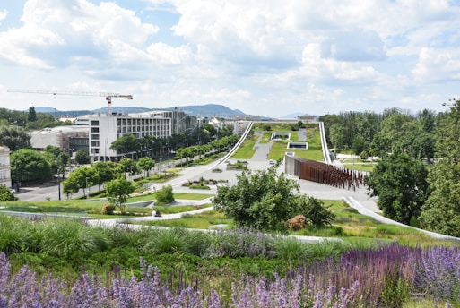 A modern landscape with expansive green spaces, pathways, and architectural elements in the foreground. Trees are spaced along the path leading up to a large, flat building in the distance. The area is lush with greenery and features both vibrant purple flowers and neatly trimmed grass. A few people are visible, enjoying the park area. A construction crane and urban buildings are visible in the background, with hills further beyond.