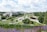 A modern landscape with expansive green spaces, pathways, and architectural elements in the foreground. Trees are spaced along the path leading up to a large, flat building in the distance. The area is lush with greenery and features both vibrant purple flowers and neatly trimmed grass. A few people are visible, enjoying the park area. A construction crane and urban buildings are visible in the background, with hills further beyond.