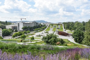A modern landscape with expansive green spaces, pathways, and architectural elements in the foreground. Trees are spaced along the path leading up to a large, flat building in the distance. The area is lush with greenery and features both vibrant purple flowers and neatly trimmed grass. A few people are visible, enjoying the park area. A construction crane and urban buildings are visible in the background, with hills further beyond.