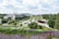 A modern landscape with expansive green spaces, pathways, and architectural elements in the foreground. Trees are spaced along the path leading up to a large, flat building in the distance. The area is lush with greenery and features both vibrant purple flowers and neatly trimmed grass. A few people are visible, enjoying the park area. A construction crane and urban buildings are visible in the background, with hills further beyond.