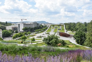 A modern landscape with expansive green spaces, pathways, and architectural elements in the foreground. Trees are spaced along the path leading up to a large, flat building in the distance. The area is lush with greenery and features both vibrant purple flowers and neatly trimmed grass. A few people are visible, enjoying the park area. A construction crane and urban buildings are visible in the background, with hills further beyond.