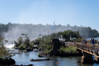 A scenic view with a bridge stretching across a river, surrounded by lush greenery. In the background, a waterfall creates mist that rises into the air. There are people walking along the bridge, and an Argentine flag is prominently displayed. The sky is clear and blue.