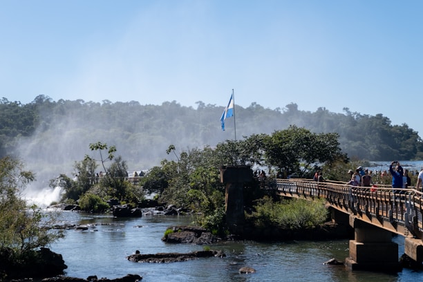 A scenic view with a bridge stretching across a river, surrounded by lush greenery. In the background, a waterfall creates mist that rises into the air. There are people walking along the bridge, and an Argentine flag is prominently displayed. The sky is clear and blue.