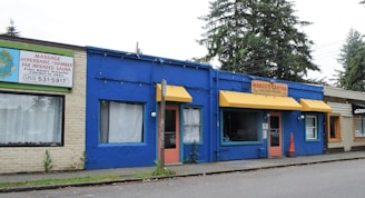 A row of small commercial buildings includes a spa with a brick exterior and a Mexican restaurant with a bright blue facade and yellow awnings. The spa advertises services such as massage and sauna treatments. There are large windows, a few of which have curtains, and a traffic cone is placed near the restaurant entrance. Tall trees are visible in the background.