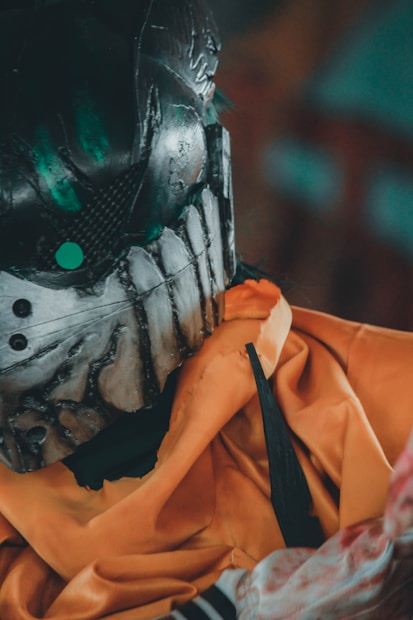 A close-up view of a person wearing a detailed, dark and metallic helmet with an intricate design and green highlights. The person is dressed in an orange garment, possibly a robe, with a layered fabric texture. The helmet has a futuristic or sci-fi aesthetic with visible rivets and textures.