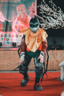 A person wearing an elaborate costume that includes a helmet with glowing green eyes, vibrant red armor, and a layered yellow and orange cape. The background features a large poster of an anime-style character alongside a tree with white flowers and a sound speaker.