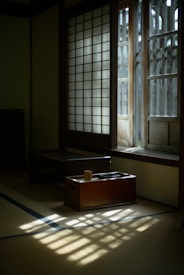 Soft sunlight filters through traditional shoji screens, casting patterned shadows on the tatami floor. A small wooden table with a cup and a book sits in the quiet, minimalist room, creating a serene and contemplative atmosphere.