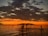 Silhouettes of people paddleboarding on calm waters beneath a dramatic sunset sky. Dark clouds add contrast to the vibrant orange and yellow hues on the horizon, while a distant island is visible.