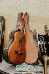 A classical guitar resting inside an open guitar case, leaning against a textured yellow wall. Alongside the guitar case is a brass trumpet, a portion of a musical keyboard, and a green and white Mexican license plate with the text 'DF MEX'. The scene appears to be a collection of musical instruments and related items.