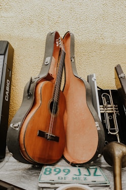 A classical guitar resting inside an open guitar case, leaning against a textured yellow wall. Alongside the guitar case is a brass trumpet, a portion of a musical keyboard, and a green and white Mexican license plate with the text 'DF MEX'. The scene appears to be a collection of musical instruments and related items.