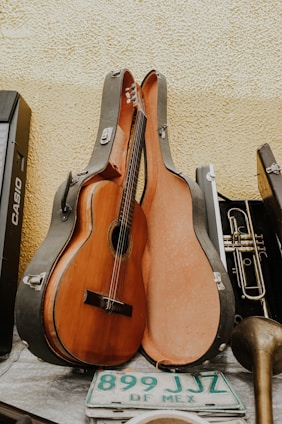 A classical guitar resting inside an open guitar case, leaning against a textured yellow wall. Alongside the guitar case is a brass trumpet, a portion of a musical keyboard, and a green and white Mexican license plate with the text 'DF MEX'. The scene appears to be a collection of musical instruments and related items.