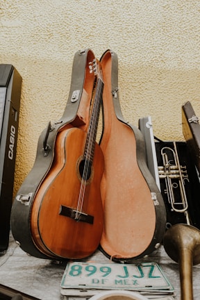 A classical guitar resting inside an open guitar case, leaning against a textured yellow wall. Alongside the guitar case is a brass trumpet, a portion of a musical keyboard, and a green and white Mexican license plate with the text 'DF MEX'. The scene appears to be a collection of musical instruments and related items.