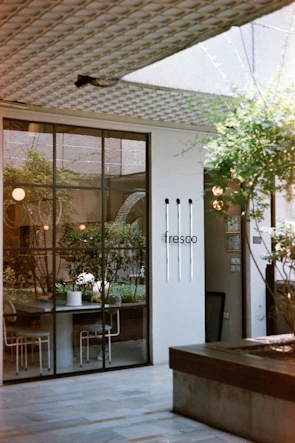 A modern interior space featuring a large window displaying an outdoor garden with greenery. The window reflects light from spherical lamps, and there are chairs and a table visible inside. A minimalist aesthetic is conveyed through the neutral color palette and clean architectural lines. The wall has the word 'fresco' and thin vertical elements adding to the design.