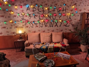 The room has retro floral wallpaper and a wooden paneling accent, featuring a sofa with a colorful crocheted blanket and a table lamp with a fringed shade. Christmas lights are strung in front of the wall, illuminating the alphabet drawn in large letters. A coffee table displays scattered colored pencils, paper, and small model figures, contributing to a cozy yet somewhat mysterious atmosphere.