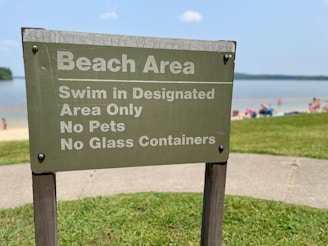 A sign at a beach area indicating rules such as swimming in designated areas only, no pets, and no glass containers. The background shows a lake or ocean, with people relaxing on a sandy shore and a grassy area nearby.