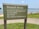 A sign at a beach area indicating rules such as swimming in designated areas only, no pets, and no glass containers. The background shows a lake or ocean, with people relaxing on a sandy shore and a grassy area nearby.