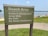 A sign at a beach area indicating rules such as swimming in designated areas only, no pets, and no glass containers. The background shows a lake or ocean, with people relaxing on a sandy shore and a grassy area nearby.