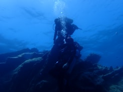 Two scuba divers exploring underwater terrain, with one diver slightly above the other. The scene is dominated by blue hues, and the divers are surrounded by rocky formations and floating bubbles.