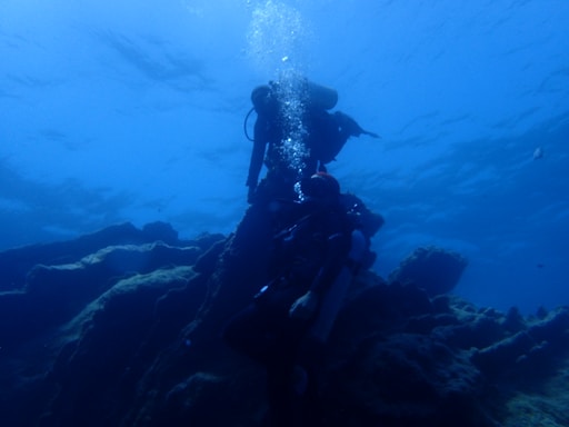 Two scuba divers exploring underwater terrain, with one diver slightly above the other. The scene is dominated by blue hues, and the divers are surrounded by rocky formations and floating bubbles.
