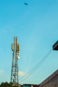 A tall communication tower stands against a bright blue sky. A bird flies near the top of the image, adding a sense of scale. The tower has multiple antennas and satellite dishes attached. Several power lines are visible, leading towards a building with a patterned wall on the right.