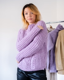 A person with blonde hair is wearing a thick, knitted lavender sweater. Their arms are folded comfortably across their body. Behind them is a clothing rack with several pieces of clothing, including a jacket and more sweaters. The setting appears to be indoors with a plain, light-colored background.