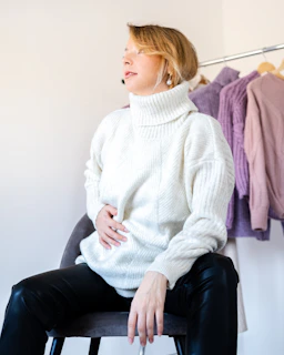 A person is seated on a chair wearing a white turtleneck sweater and black pants. Their hand rests on their abdomen, and they appear to be looking upward. In the background, there are several pastel-colored sweaters hanging on a clothing rack.