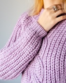 A close-up of a person's hand resting on a lavender-colored, chunky knit sweater. The hand is adorned with a large, ornate ring featuring a prominent gemstone. The sweater features a thick, textured pattern, and the person's blonde hair is partially visible above.