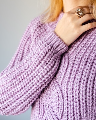 A close-up of a person's hand resting on a lavender-colored, chunky knit sweater. The hand is adorned with a large, ornate ring featuring a prominent gemstone. The sweater features a thick, textured pattern, and the person's blonde hair is partially visible above.