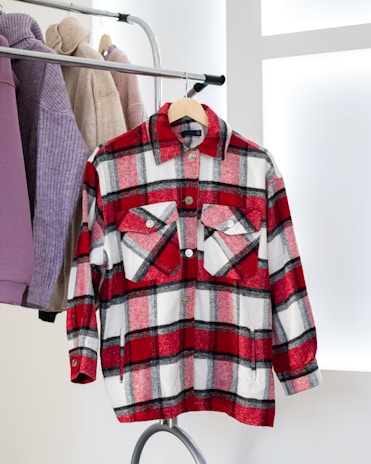 A red and white plaid button-up shirt is hanging on a clothes rack. Beside it are other garments in neutral tones like beige and purple. The setting is bright with a modern, minimalistic design.