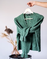 A hand holds a hanger displaying a green knitted cardigan. The background features a minimalist scene with a vase filled with dried grasses sitting on a small black table, creating a cozy and natural ambiance.