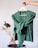 A hand holds a hanger displaying a green knitted cardigan. The background features a minimalist scene with a vase filled with dried grasses sitting on a small black table, creating a cozy and natural ambiance.