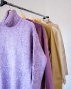 A set of sweaters hanging on wooden hangers on a metal clothing rack. The sweaters are in different colors, including purple, beige, and tan, and are made of a thick, warm material. The focus is on the textures and colors of the knitwear.