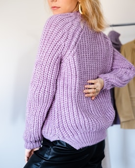 A person wearing a thick, textured purple sweater, turned slightly away, with a hand on their hip. The sweater's knit is prominent, and the person has blonde hair with a gold earring visible. In the background, blurred clothing can be seen on hangers.