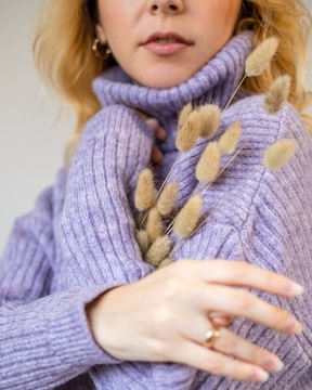 A person wearing a cozy, textured lavender sweater holds delicate, fluffy dried grasses. The close-up focus highlights the softness of the fabric and the natural elements. The person's hand is gently wrapped around the bouquet, conveying warmth and comfort.