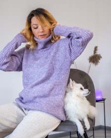 A person with blonde hair is seated on a chair wearing a cozy, purple turtleneck sweater. A white fluffy dog is comfortably sitting next to them, appearing relaxed. The background features a neutral-colored wall with some decorative pampas grass and a soft glowing light, creating a warm ambiance.