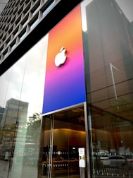 A modern building facade featuring a prominent Apple logo on a gradient background with colors transitioning from blue to pink and orange. The building has large glass windows revealing the interior of what appears to be an Apple store, with wooden tables and electronic products. Reflections of the city skyline are visible in the glass.