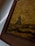 A close-up view of a framed artwork with earthy textures. The image features a rustic landscape depiction with a brownish gold hue dominating the canvas. The artwork shows abstract shapes that resemble a natural scene, possibly with a focus on earthy or sandy terrain.