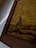 A close-up view of a framed artwork with earthy textures. The image features a rustic landscape depiction with a brownish gold hue dominating the canvas. The artwork shows abstract shapes that resemble a natural scene, possibly with a focus on earthy or sandy terrain.