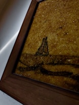 A close-up view of a framed artwork with earthy textures. The image features a rustic landscape depiction with a brownish gold hue dominating the canvas. The artwork shows abstract shapes that resemble a natural scene, possibly with a focus on earthy or sandy terrain.
