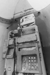A vintage public telephone with a keypad and handset. The device is weathered and shows signs of rust and neglect. It is located in an enclosed booth with graffiti and peeling paint on the surrounding walls.