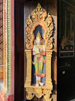 A decorative statue set within an ornate alcove, featuring a figure adorned with intricate details and vibrant colors. The alcove is richly carved with floral and ornamental patterns, highlighted by a bright yellow and red color scheme.